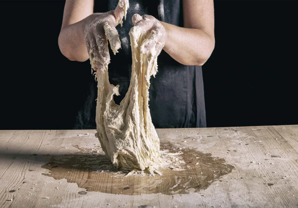 Mani infarinate che tirano e lavorano l’impasto del pane su un tavolo di legno, con un grembiule scuro sullo sfondo nero