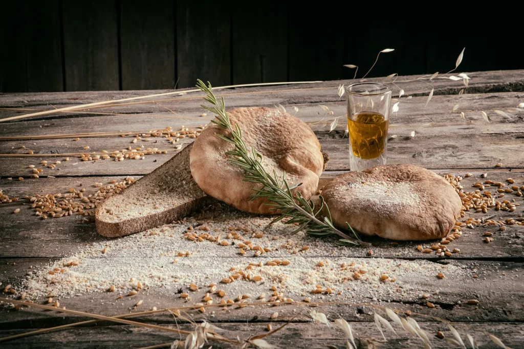 Pane Nostro: pagnotte di pane casereccio e una fetta di pane, chicchi di grano, rametto di rosmarino e un bicchiere d’olio su un vecchio tavolo di legno