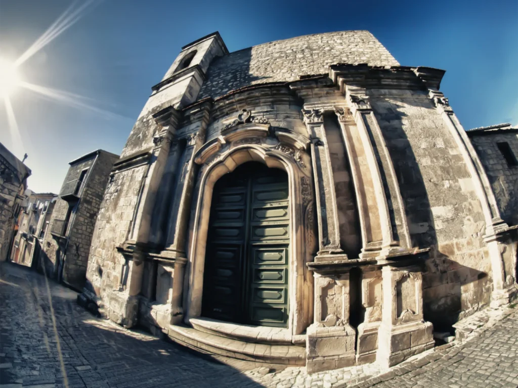 Facciata in pietra della Chiesa del S. Salvatore a Petralia Soprana, con il portale barocco e il grande portone verde, sotto il sole