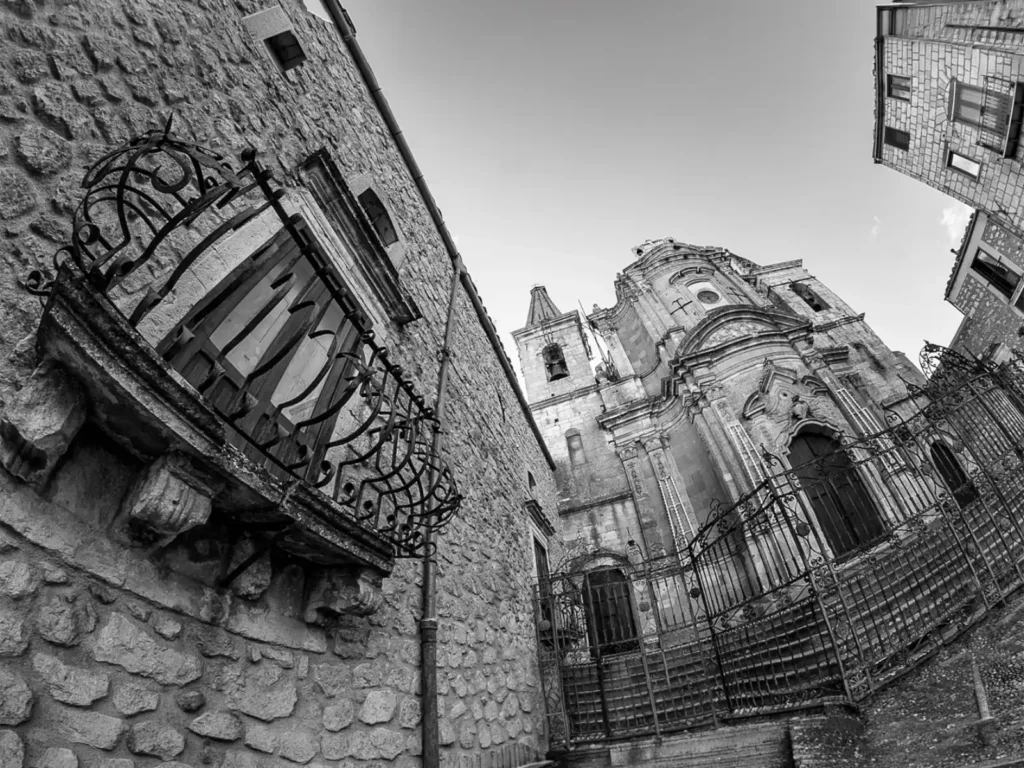 Chiesa di S. Maria di Loreto a Petralia Soprana con la cancellata in ferro battuto e, in primo piano, un balcone in ferro su una casa in pietra, in bianco e nero