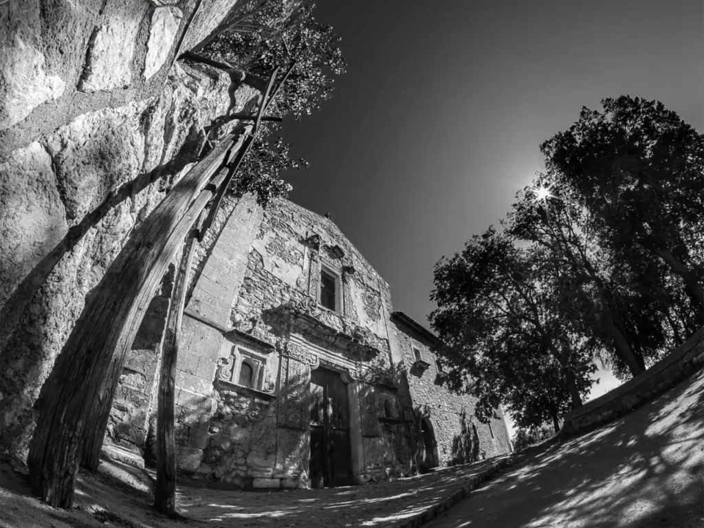 Chiesa del Convento dei Frati Minori Riformati a Petralia Soprana, con il portale barocco in pietra tra una parete di roccia e grandi alberi, in bianco e nero