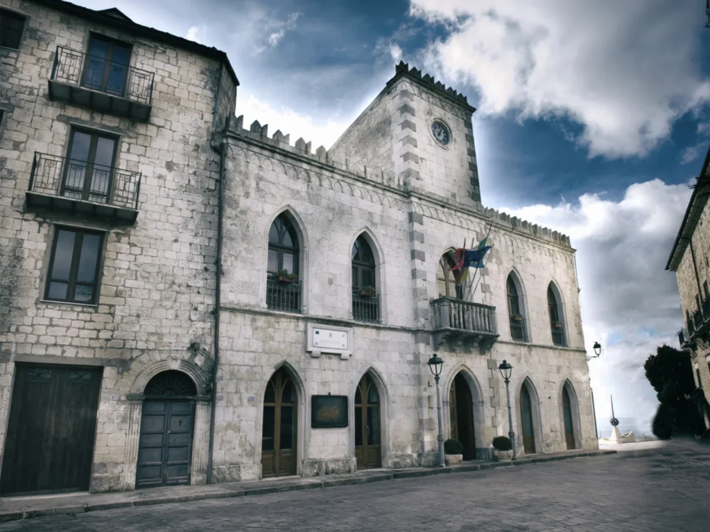 Palazzo del Municipio di Petralia Soprana in pietra chiara, con finestre ad arco acuto, merli, torre dell’orologio e bandiere sul balcone