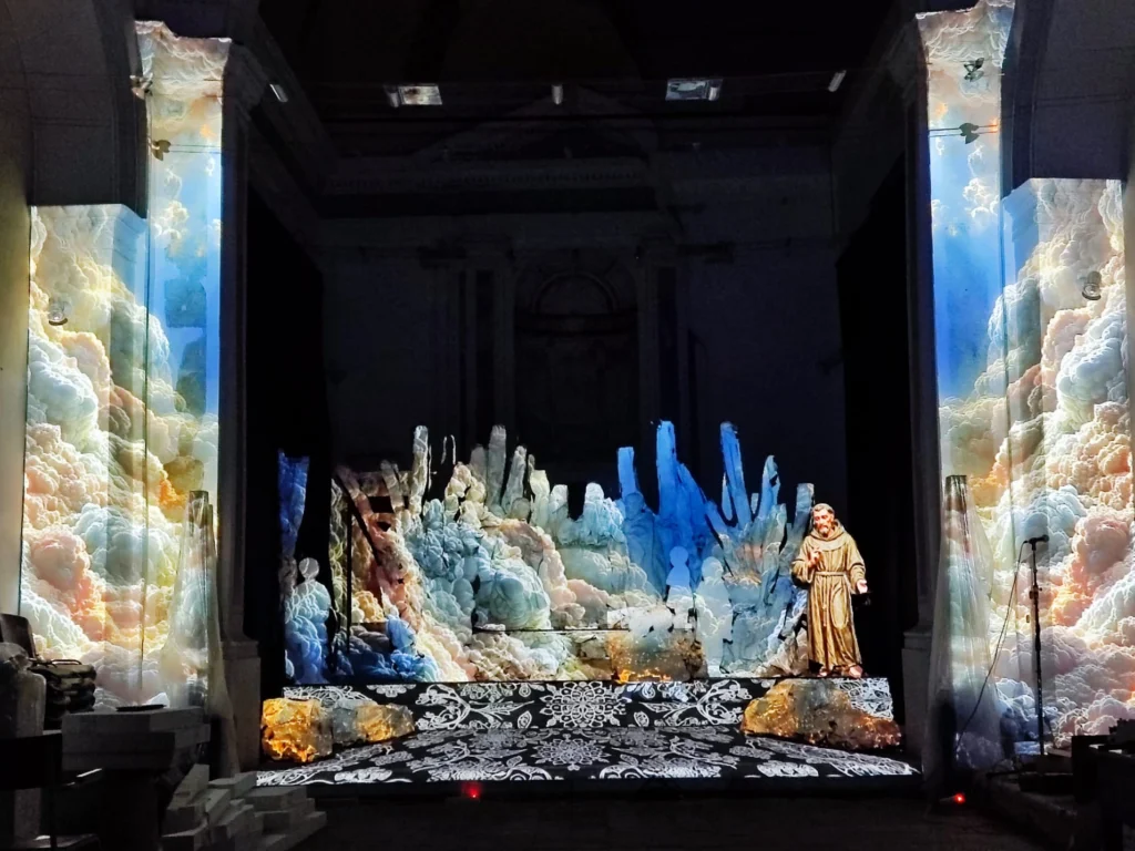 Interno della chiesa con nuvole colorate proiettate dal video mapping su colonne e scenografia, pavimento a merletto e statua di San Francesco, Presepe d’InCanto