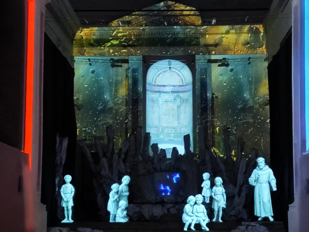 Statue bianche di bambini e di San Francesco davanti a una grotta scura, con l’abside illuminata dal video mapping del Presepe d’InCanto